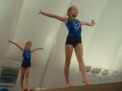 Twee jonge turnsters in blauwe turnpakken oefenen op een evenwichtsbalk in een gymzaal.