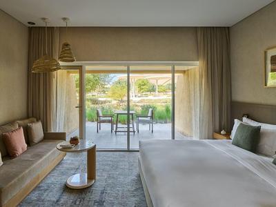 Modernes Hotelzimmer mit großem Bett, Sofa, Teppich und bodentiefen Fenstern zum Garten.