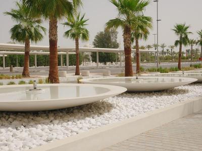 Moderne Parkanlage mit runden Wasserbecken, weißen Kieselsteinen und hohen Palmen am Gehweg.