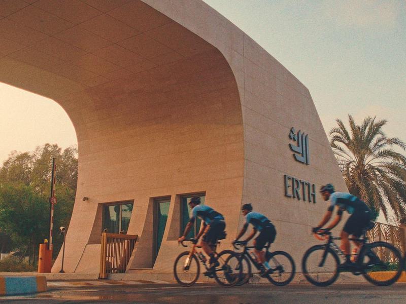 Drei Radfahrer fahren an einem großen beige-farbenen Bogen mit Palmen im Hintergrund vorbei.