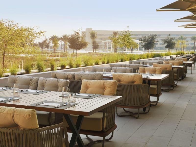 Leere Außenterrasse mit gestuften Tischen, Polstersesseln und Blick auf grüne Bäume und Gebäude.