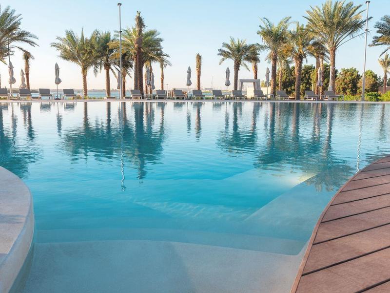 Klares Schwimmbecken mit Palmen und Sonnenschirmen unter blauem Himmel am Ufer.