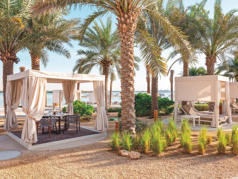 Strand mit Palmen, beigen Zelten und Sitzgelegenheiten bei Sonnenschein und klarem Himmel.