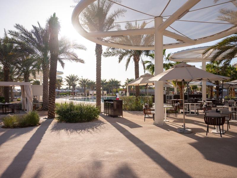 Sonniger Außenbereich mit Palmen, Sitzgruppen, Sonnenschirmen und Schatten durch Pergola.