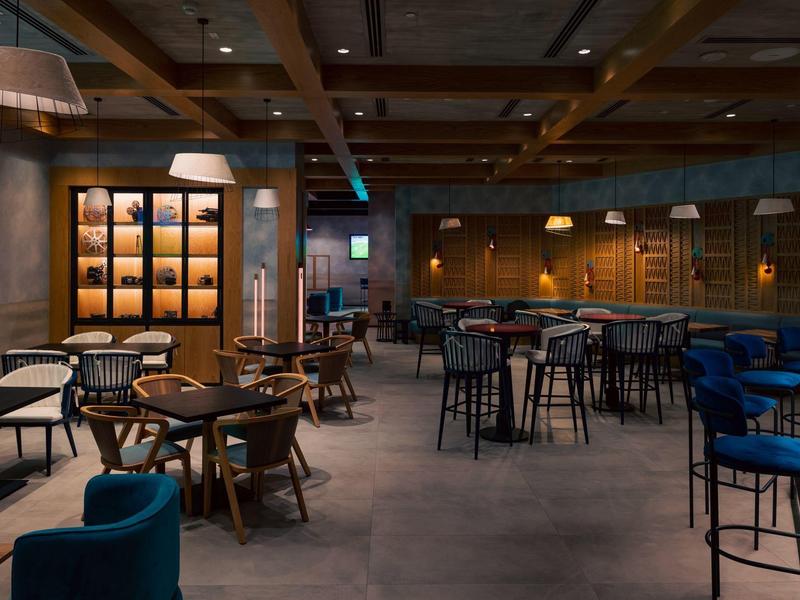 Moderne Bar mit Holztischen, blauen Sesseln, Barhockern und warmem Licht im Innenraum.