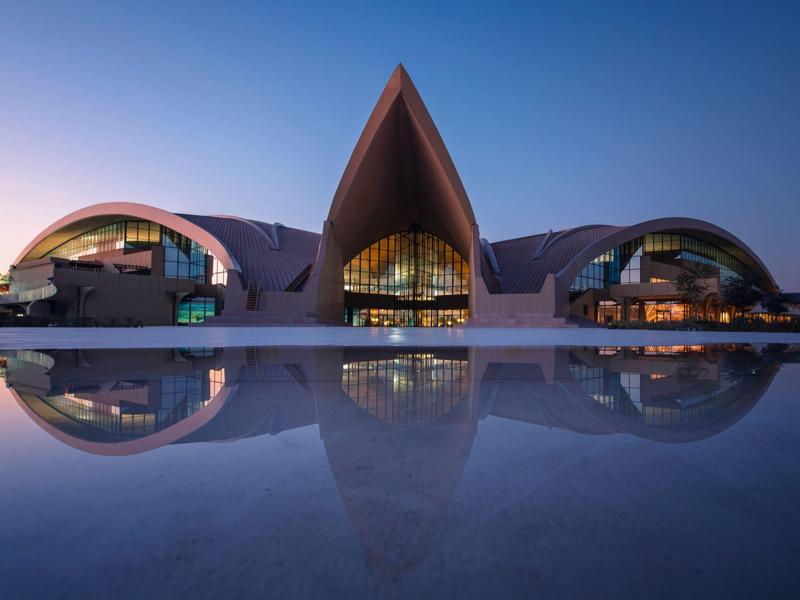 Modernes Gebäude mit spitzem Dach, gebogenen Seiten und Spiegelung im Wasser bei Dämmerung.