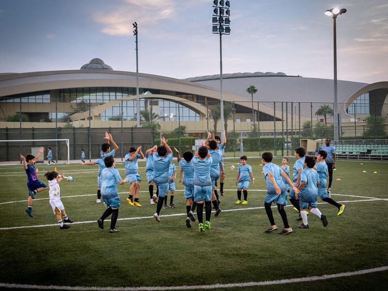 Kinder in hellblauen Trikots jubeln auf dem Fußballfeld vor einem modernen, großen Stadion.