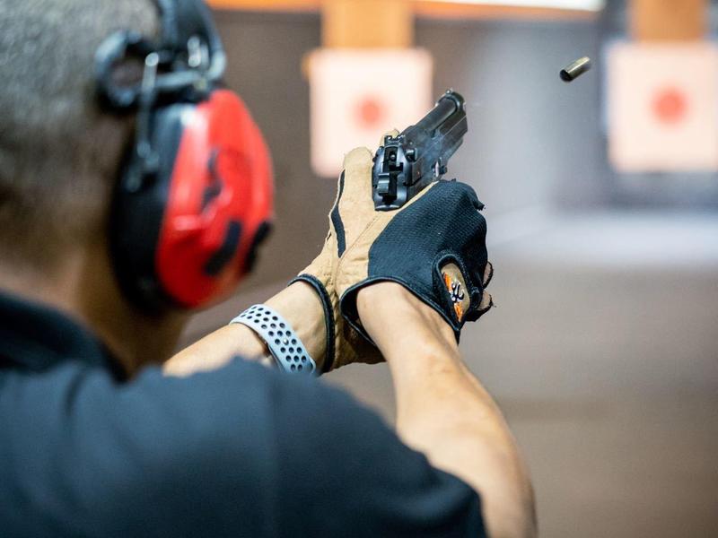 Mann mit Schutzbrille und Kopfhörern schießt Pistole auf Schießstand, aufgeblasenem Patronenhülse sichtbar.