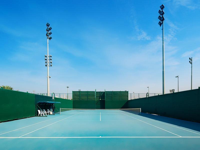 Leerer Tennisplatz mit blauem Spielfeld, grünem Zaun und hohen Flutlichtmasten bei klarem Himmel.
