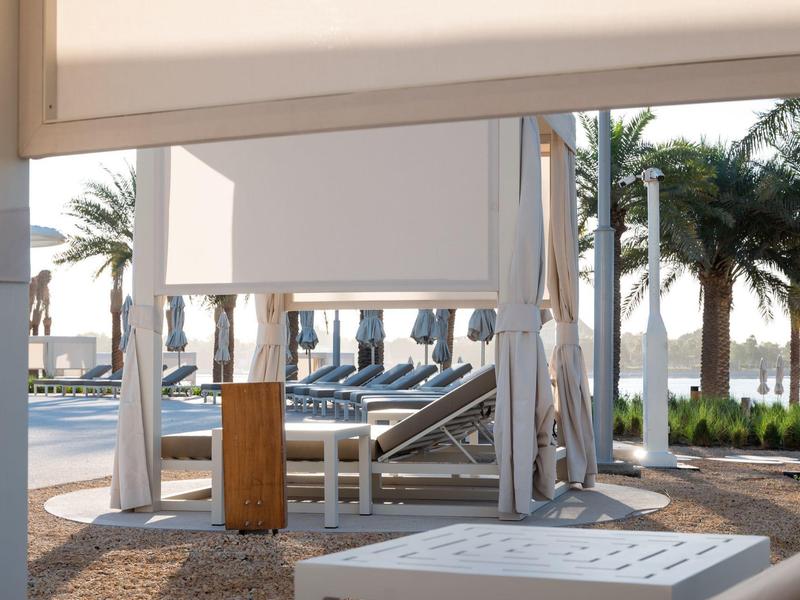 Moderner Strandpavillon mit weißen Vorhängen, Liegestühlen und Palmen im Hintergrund bei Sonnenlicht.