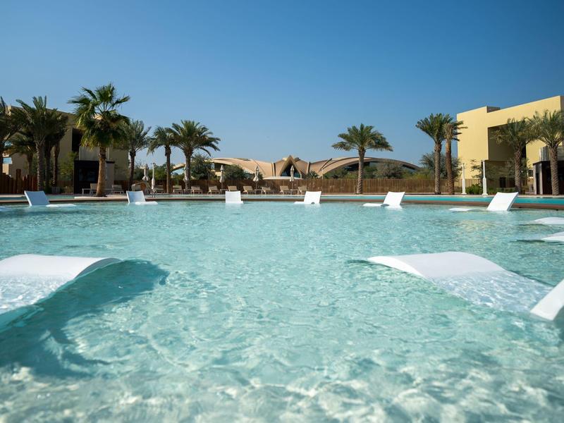 Großer Pool mit klar blauem Wasser, weißen Liegen im Wasser und Palmen im Hintergrund unter blauem Himmel.