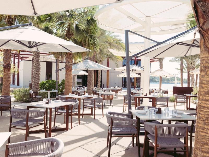 Outdoor-Café mit Tischen, Stühlen und großen Sonnenschirmen, umgeben von Palmen.