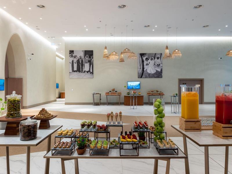 Moderner Frühstücksraum mit Buffet, weißen Wänden, Holztischen und bunten Getränken am Buffet.