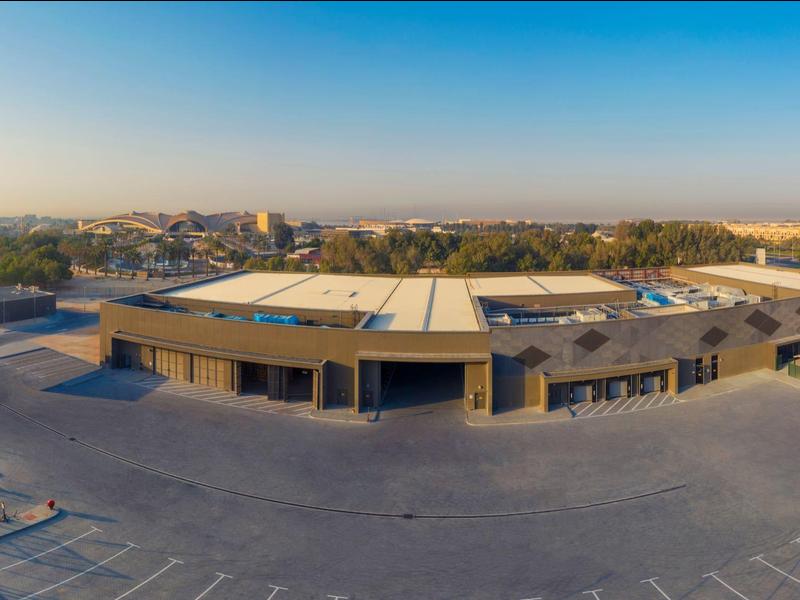 Großes Industriegebäude mit Parkplätzen, Straße im Vordergrund, städtische Umgebung und blauer Himmel.