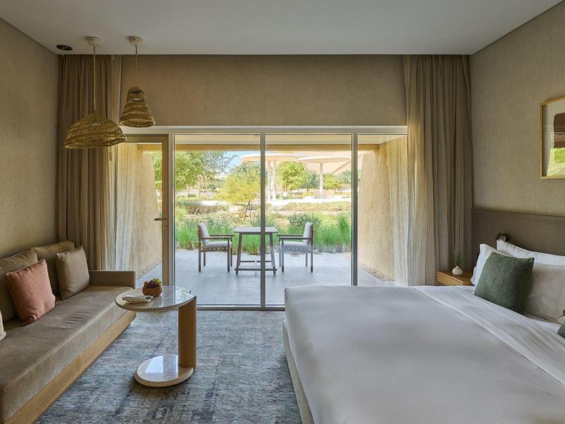 Modernes Hotelzimmer mit großem Bett, Sofa, Teppich und bodentiefen Fenstern zum Garten.