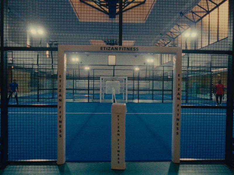 Padel-Tennisplatz mit blauem Boden, Umzäunung und beleuchteter Halle in gedämpftem Licht.