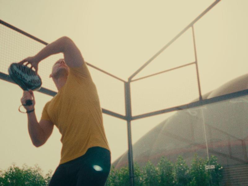 Ein Mann in gelbem T-Shirt wirft einen Baseball im sonnigen Trainingsfeld mit Zaun.