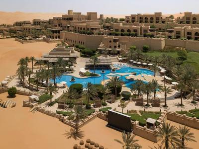 Desert resort with a large circular pool surrounded by palm trees and terraced buildings.