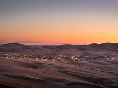 Desert camp with lights glowing at sunset across rolling sand dunes under a clear sky.