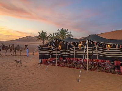 Bedouin tent setup in desert with camels, palm trees, and a dog at sunset.