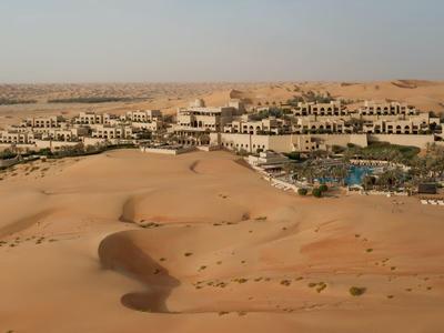 Desert resort with sandy dunes, multiple buildings, and a pool under a clear sky.