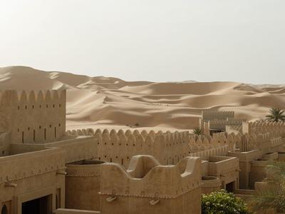 Desert fort with tan walls and palm trees against sandy dunes under a hazy sky.