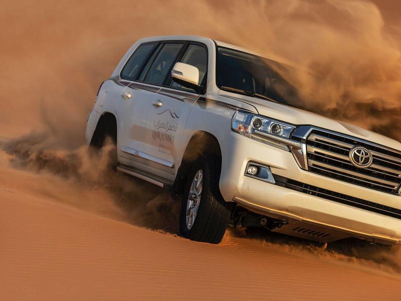 White SUV driving fast on sandy desert, creating dust clouds around it.