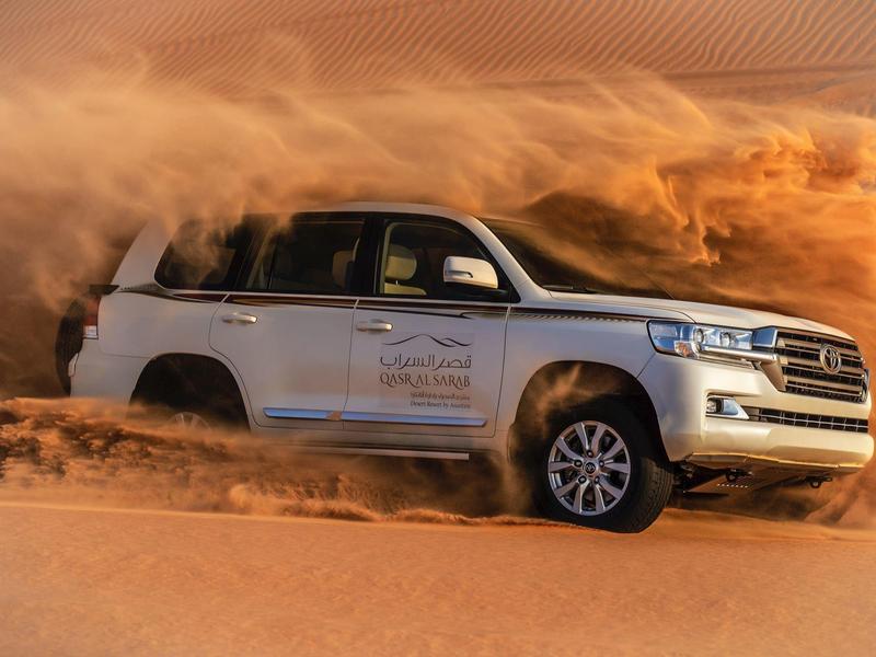 White SUV driving fast through desert sand, kicking up large sand cloud at sunset.