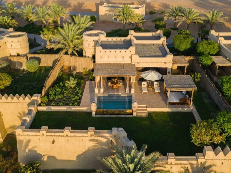 Aerial view of a desert-style hotel with a pool, palm trees, and sandy surroundings.