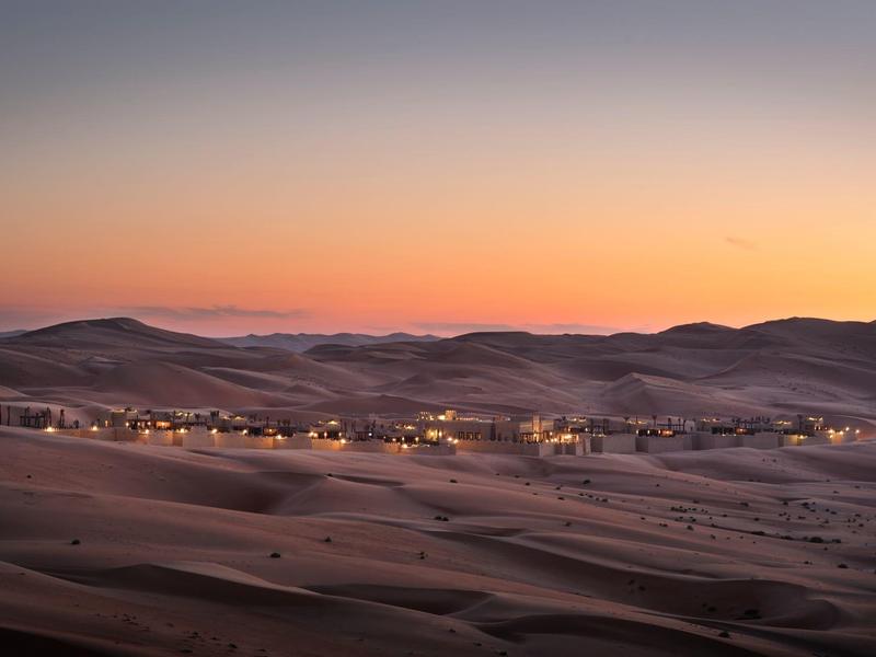 Desert camp with lights glowing at sunset across rolling sand dunes under a clear sky.