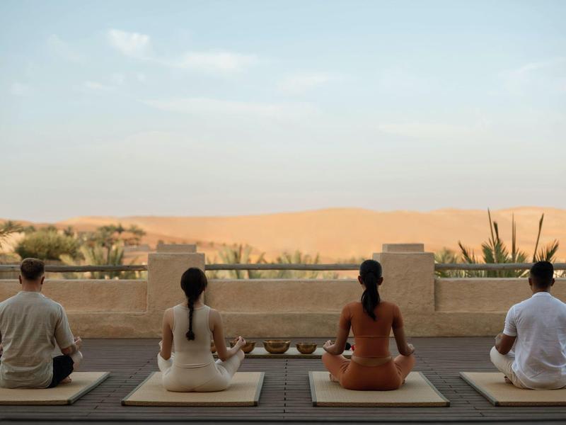 Four people meditate on cushions facing a desert view with clear sky and distant dunes.