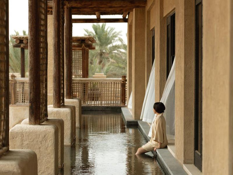 A person sitting beside a narrow reflective water canal under a wooden roofed corridor.