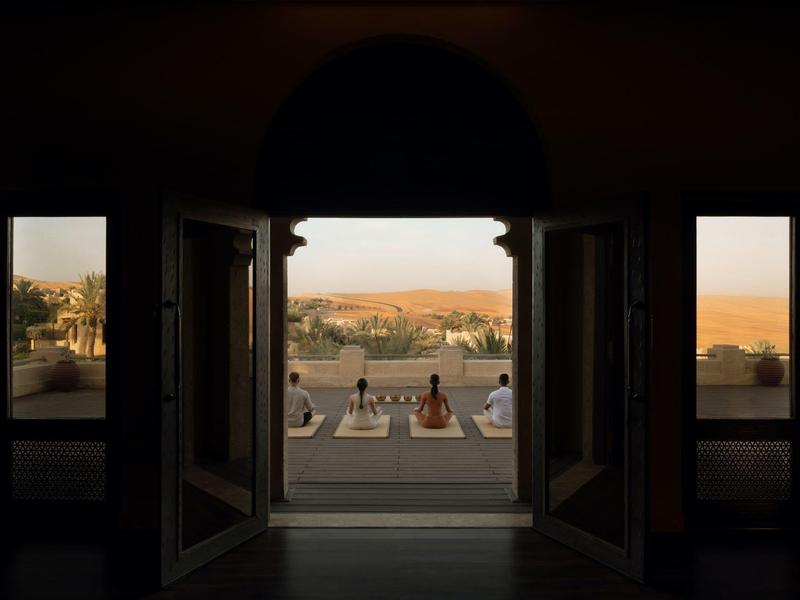 View from a dark room through open doors onto people sitting by a sunlit pool with hills in the background.