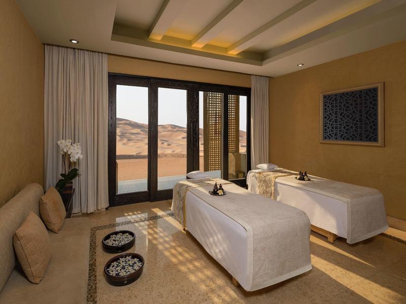 Serene spa room with two massage tables, beige walls, cushioned bench, and large windows.