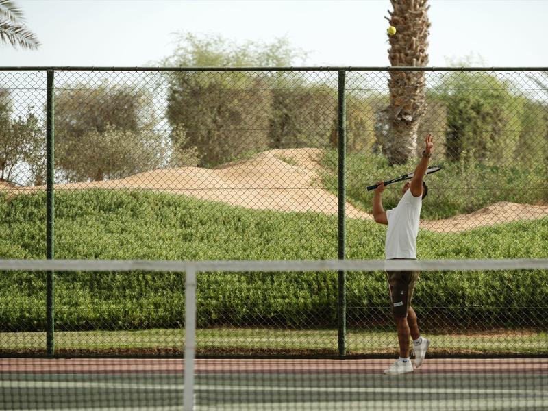 Person preparing to serve on an outdoor tennis court surrounded by trees and sand dunes.