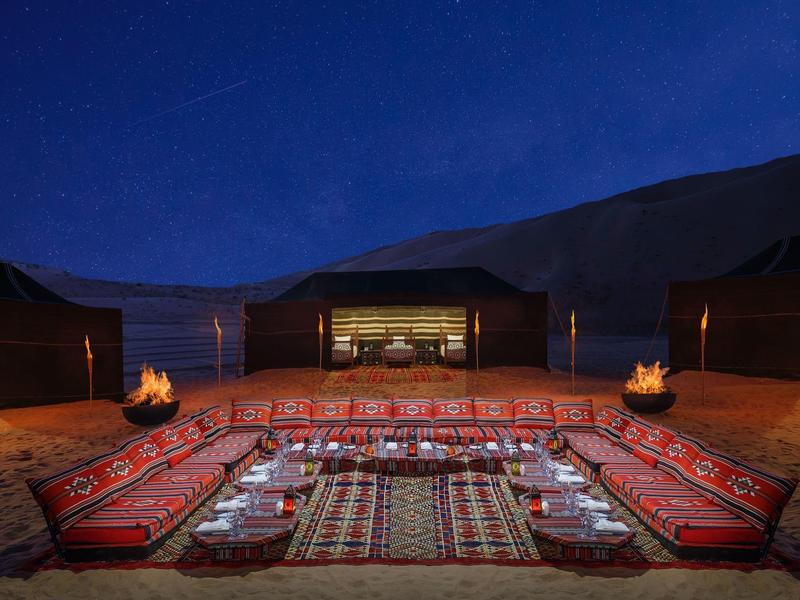 Night desert camp with cushions, rugs, tents, and fire pits under a deep blue sky.