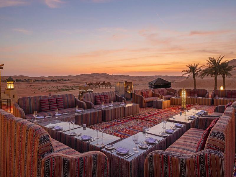Outdoor desert lounge with sofas, rugs, and dining setup at sunset under a partly cloudy sky.