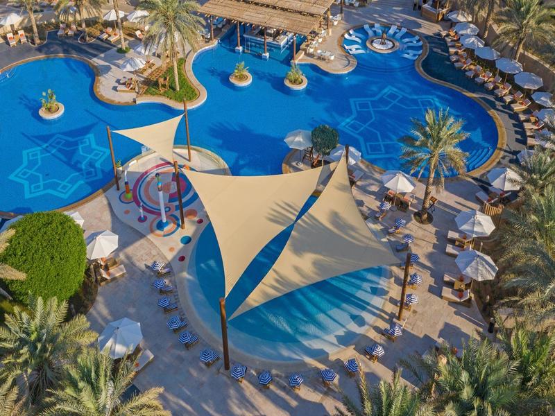 Aerial view of a resort pool area with umbrellas, lounge chairs, palm trees, and shaded seating.