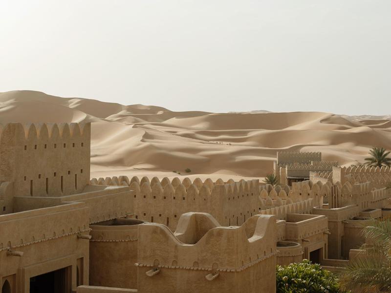Desert fort with tan walls and palm trees against sandy dunes under a hazy sky.