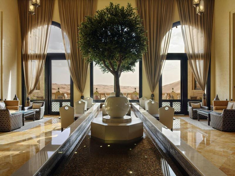 Luxurious hotel lobby with tall windows, elegant curtains, a central tree, and comfortable seating.