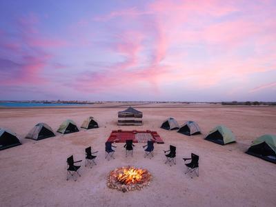 Campement de plage avec tentes et chaises autour d'un feu de camp au coucher du soleil.
