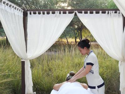 Frau massiert eine Person unter einem weißen Baldachin im Freien, umgeben von Gras und Bäumen.