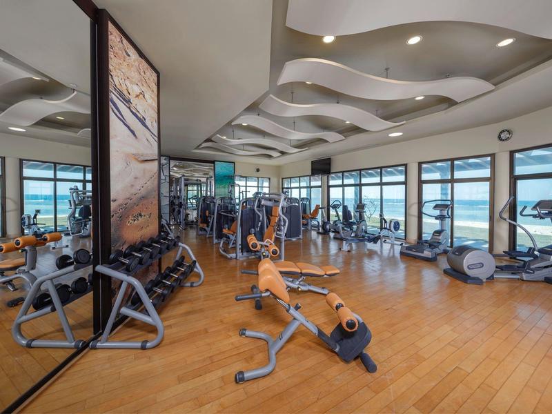 Heller Fitnessraum mit Holzfußboden, Hantelregal, Geräten und großen Fenstern mit Außenblick.