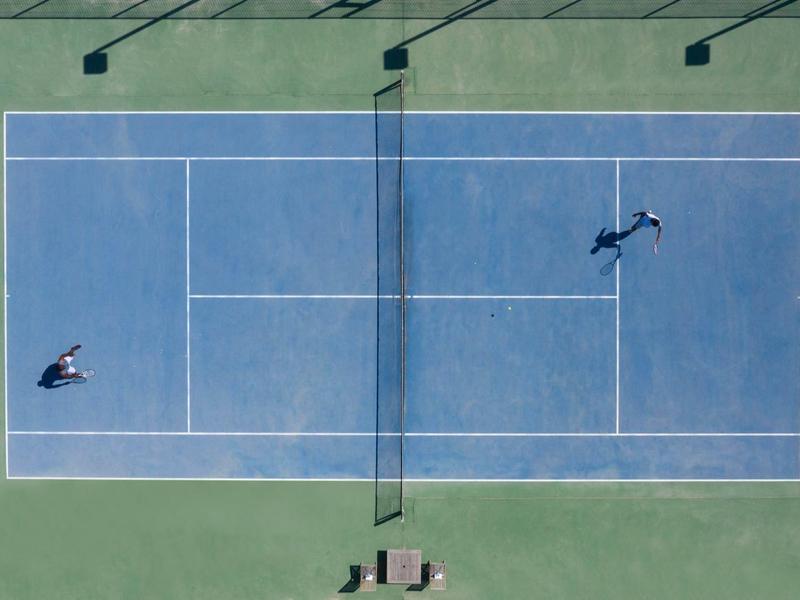 Vogelperspektive eines blauen Tennisplatzes mit zwei Spielern und Schatten der Tennismasten.