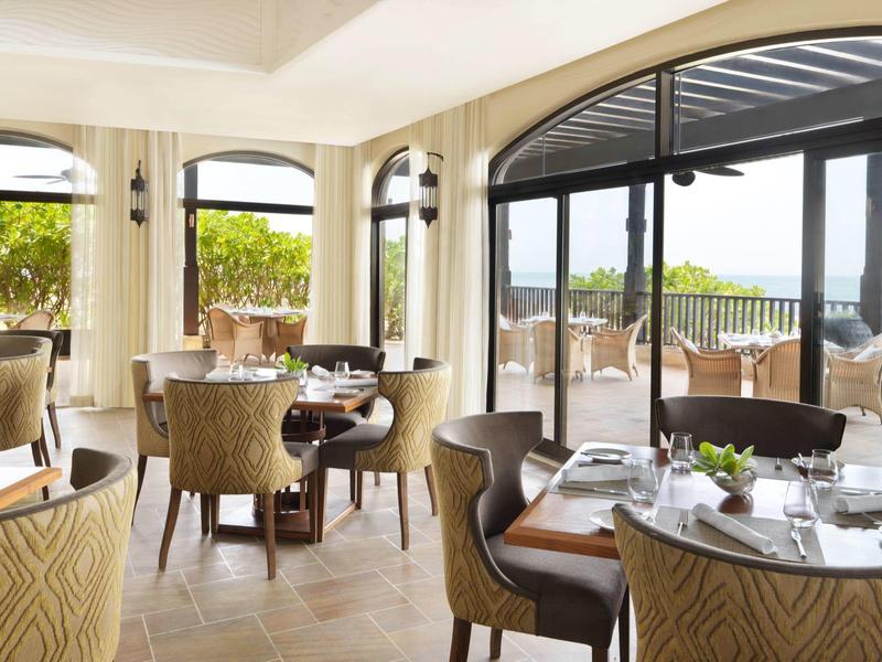 Helles Restaurant mit großen Fenstern, runden Tischen, bequemen Stühlen und Meerblick außen.