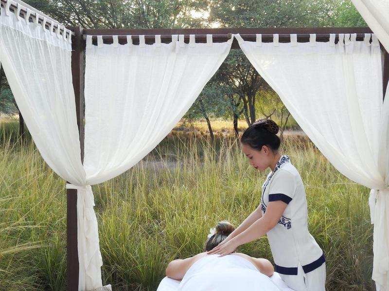 Frau massiert eine Person unter einem weißen Baldachin im Freien, umgeben von Gras und Bäumen.