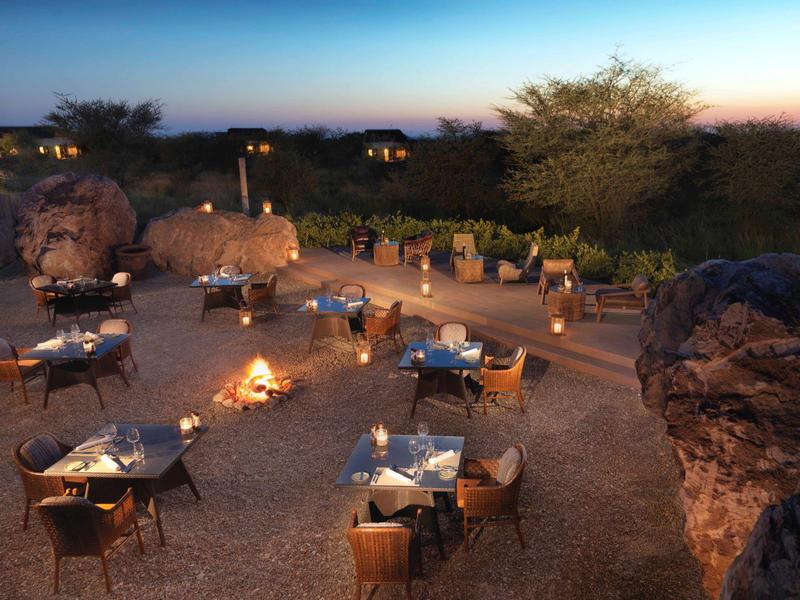 Romantisches Freiluft-Dinner mit Feuern und Tischen in einer wüstenähnlichen Landschaft bei Sonnenuntergang.