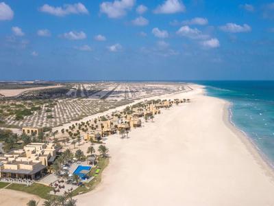 Aerial view of a large beach resort with white sand, blue pool, palm trees, and clear ocean water.
