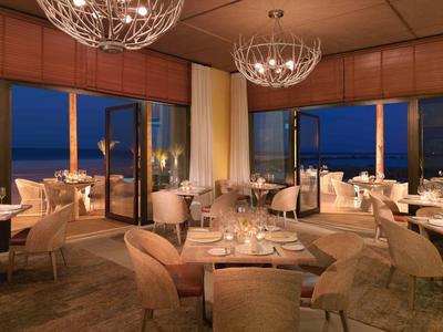 Cozy hotel restaurant at night with elegant lighting and ocean views through large windows.
