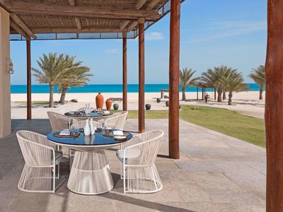 Outdoor dining area with modern chairs and table overlooking palm trees and beach by the sea.